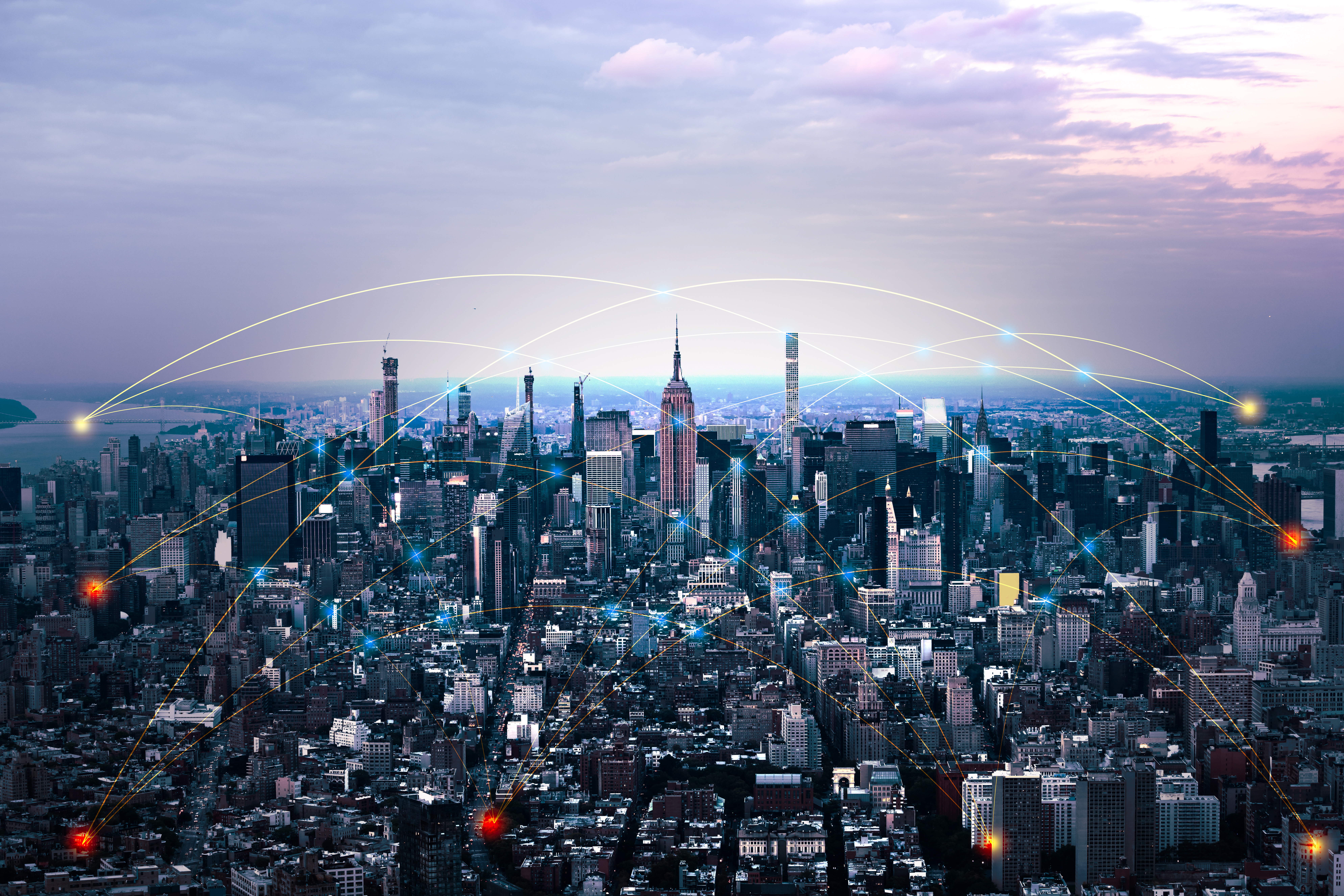 Aerial view of a dense city skyline at dusk, with skyscrapers stretching across the horizon. Glowing lines and nodes overlay the buildings, illustrating digital connectivity and data networks linking different parts of the city.