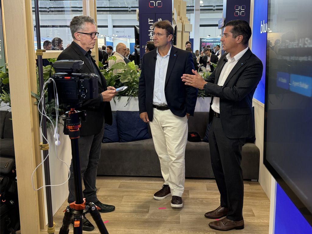 Three conference attendees participate in an on-camera discussion at the Blue Planet booth, with recording equipment visible in the foreground.