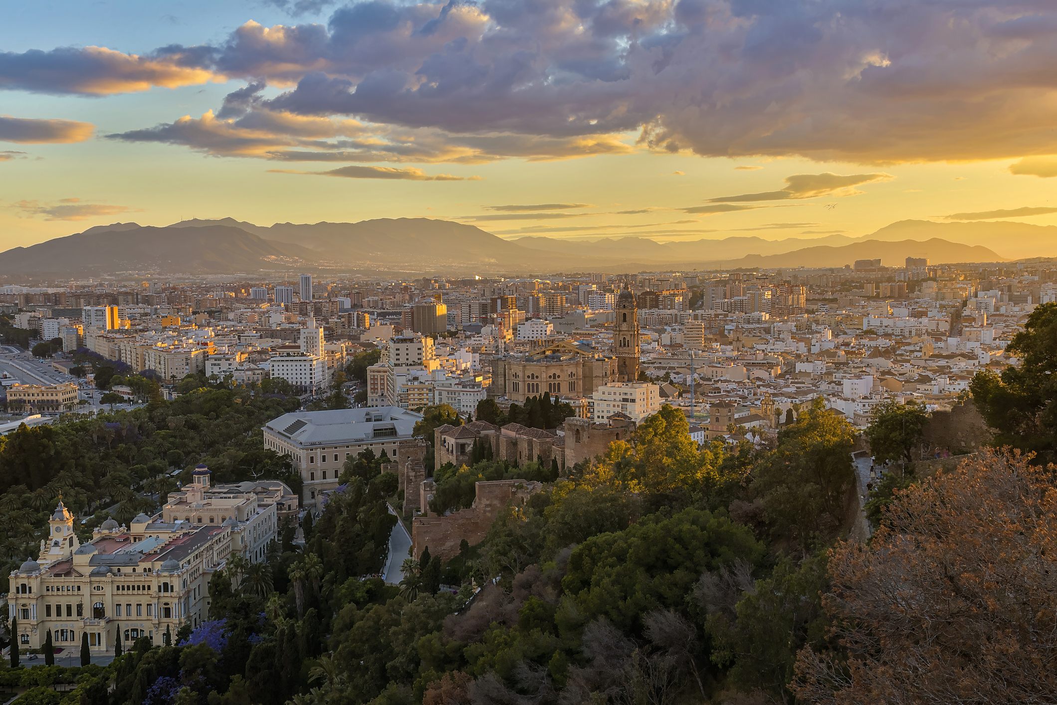 Panoramic view of Málaga, Spain, showing the city center, historic architecture, tree-covered hills, and mountain ranges illuminated by warm evening light.