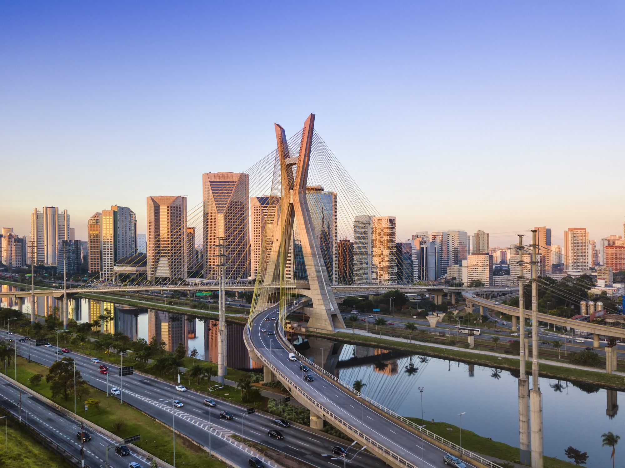 São Paulo skyline at sunset with a cable-stayed bridge, highways, and reflections along the river.