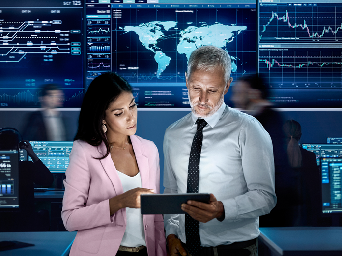 Two professionals reviewing data on a tablet in a high‑tech operations center with global analytics dashboards