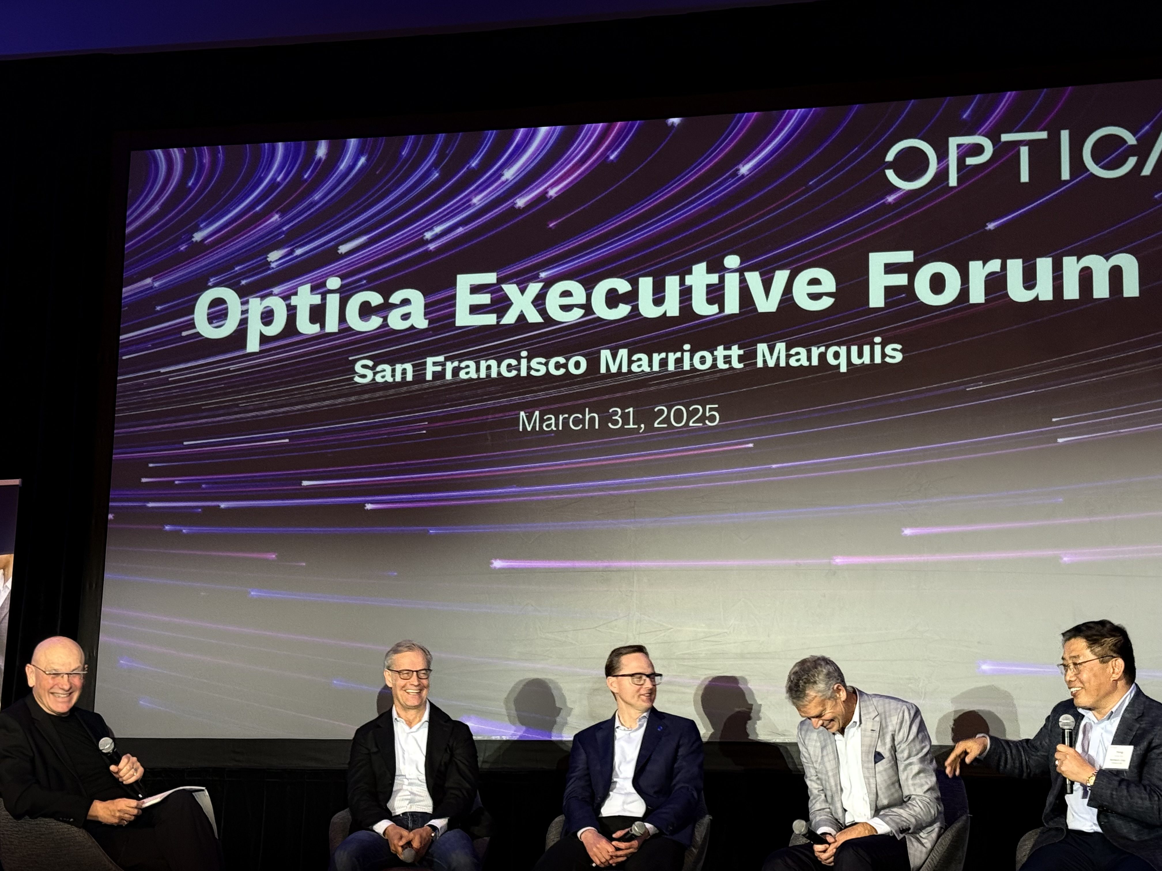 a panel of men in front of a screen on a stage at OFC
